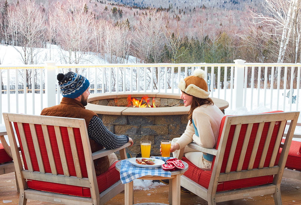 a man and woman sitting outside by a fire pit in winter with snacks and drinks