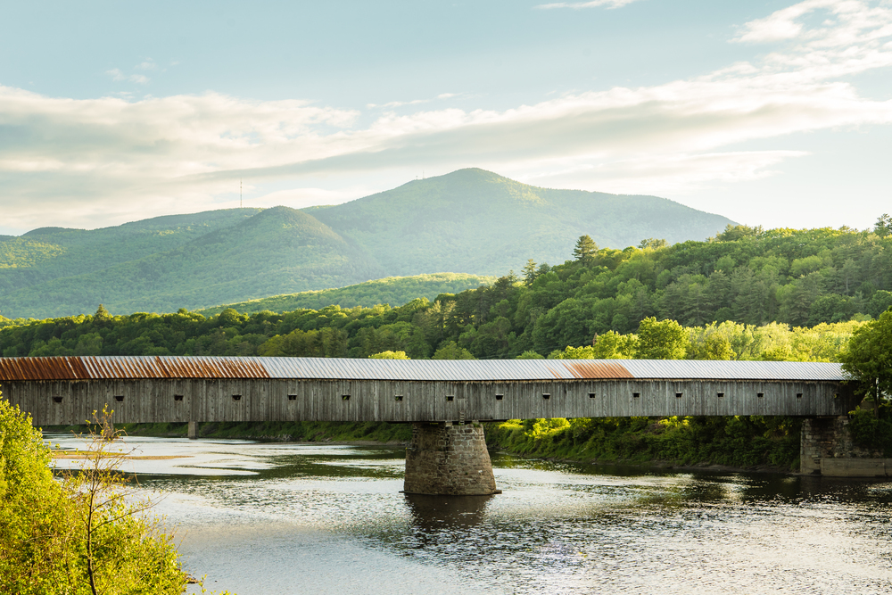 A covered bridge crossing a river with a mountain landscape behind it. 