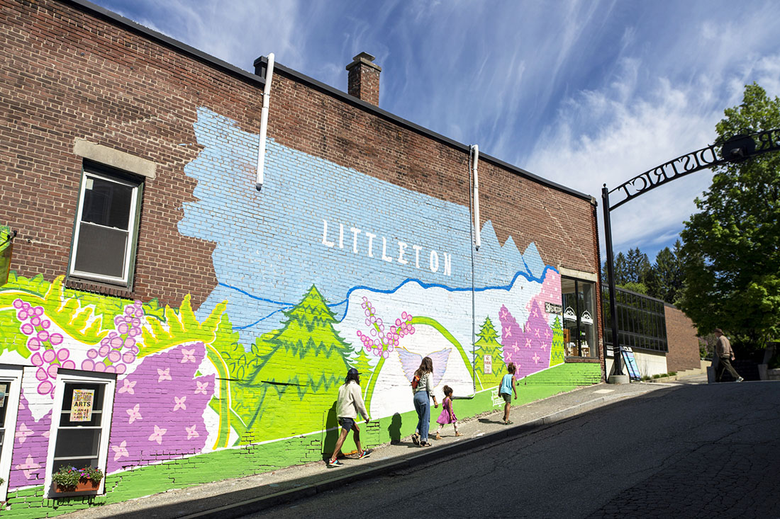 a family walking on a sidewalk up a hill next to a mural that says "Littleton"