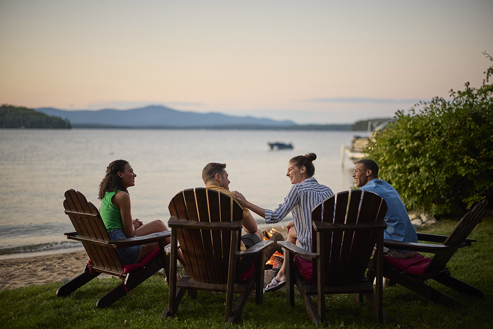 a group of people sitting at the edge of a lake at sunset by a fire pit.