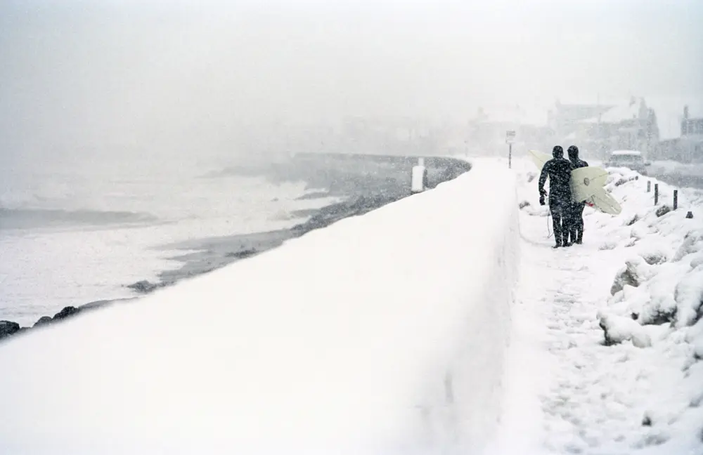 Two people walking along sea edge with surfboards during a snow storm