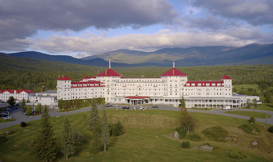 An image of a large white hotel with a red roof surrounded by mountains
