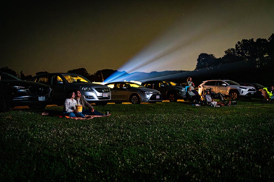 two people sitting on a lawn in front of parked cars at a drive-in theater.