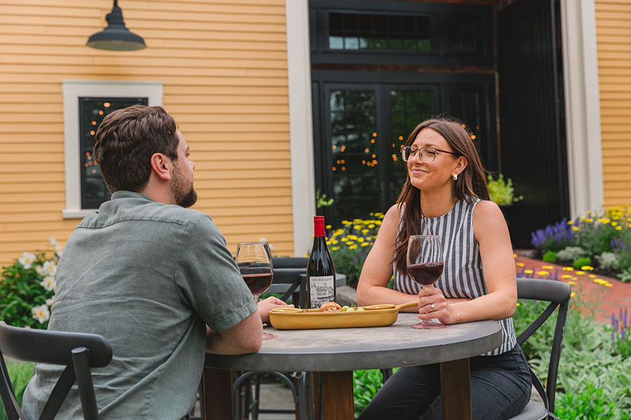 a man and woman dining outside with wine by a barn and flowers.