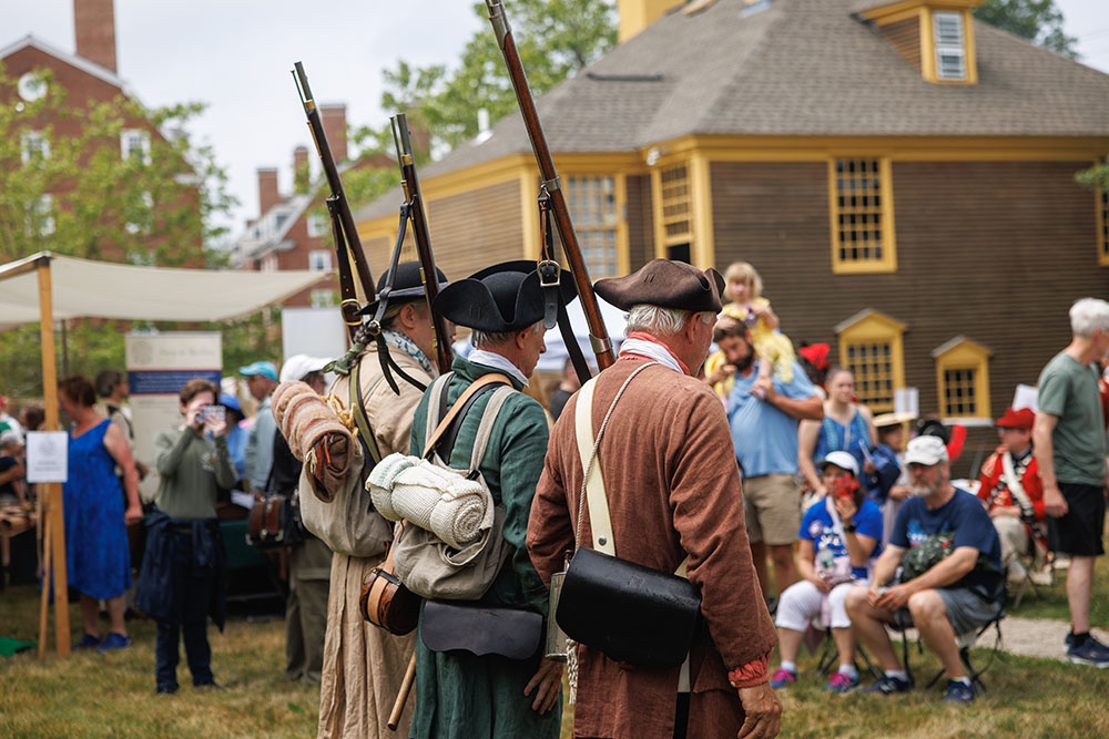 three men dressed in historical clothing outside a historical building with a crowd gathered.
